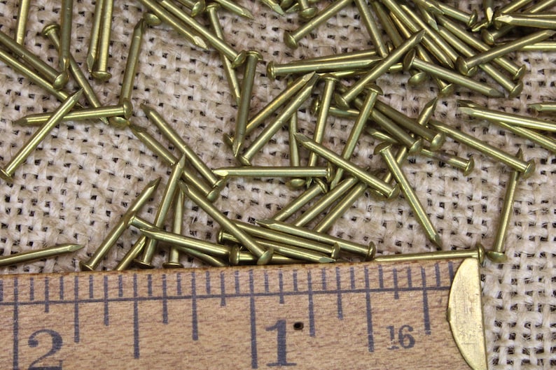May include: A close-up of a pile of brass tacks, with a wooden ruler measuring 1 inch and 2 inches. The tacks are scattered on a burlap surface.