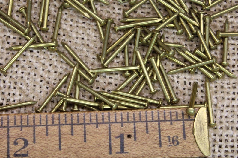 May include: A close-up of a pile of brass tacks on a burlap surface. A wooden ruler with inch markings is visible at the bottom of the image. The ruler shows measurements of 1 inch and 2 inches.
