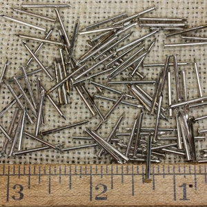 May include: A close-up of a pile of silver-colored tacks or upholstery nails on a burlap surface. A wooden ruler with inch markings is placed below the tacks.