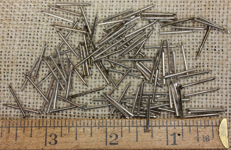May include: A close-up of a pile of silver-colored tacks or upholstery nails on a burlap surface. A wooden ruler with inch markings is placed below the tacks.