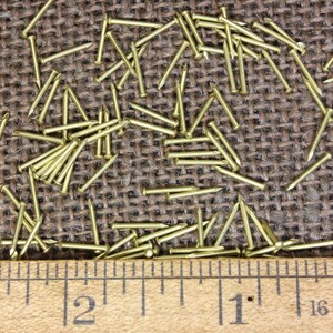 May include: A close-up of a ruler measuring 3 inches with a pile of small, gold-colored tacks or nails scattered on a brown burlap surface.