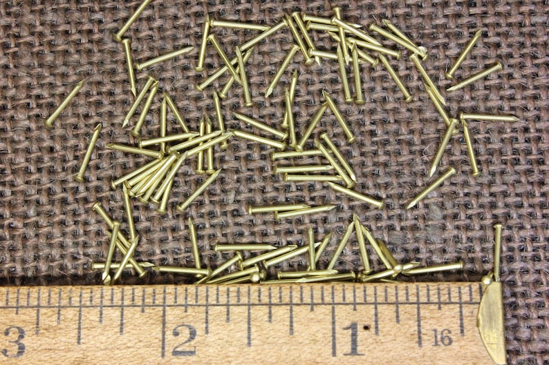May include: A close-up of a ruler measuring 3 inches with a pile of small, gold-colored tacks or nails scattered on a brown burlap surface.