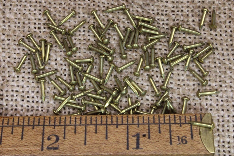 May include: A close-up of a group of small, gold-coloured tacks or nails scattered on a hessian surface. A wooden ruler with inch markings is placed below the tacks, showing a measurement of 1 inch and 2 inches.