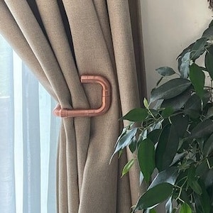 May include: A copper pipe curtain tieback holding back a pair of beige curtains. A potted plant with green leaves is in the background, next to a blue electric guitar.