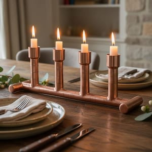 May include: A copper candle holder with four lit candles. The candle holder is made of copper pipes and sits on a wooden table. The table is set with plates, cutlery, and napkins. The background includes a window, a fireplace, and a bookshelf.
