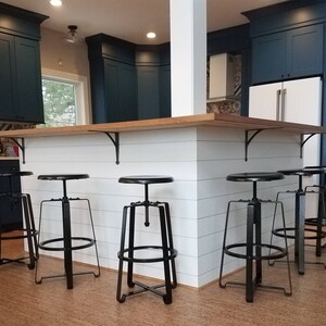 An industrial themed kitchen with navy blue kitchen cabinets, using industrial black wrought iron stools with black cast iron shelving brackets under a wood kitchen worktop with a white panelled kitchen island. Spot lights to ceiling and a lovely wooden floor.