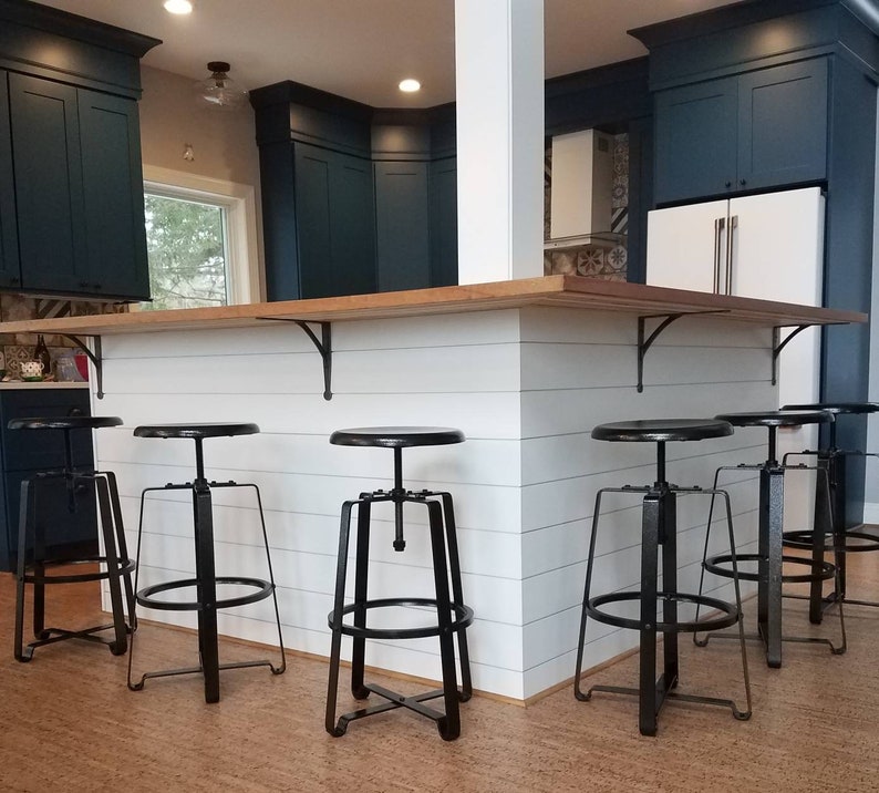 An industrial themed kitchen with navy blue kitchen cabinets, using industrial black wrought iron stools with black cast iron shelving brackets under a wood kitchen worktop with a white panelled kitchen island. Spot lights to ceiling and a lovely wooden floor.