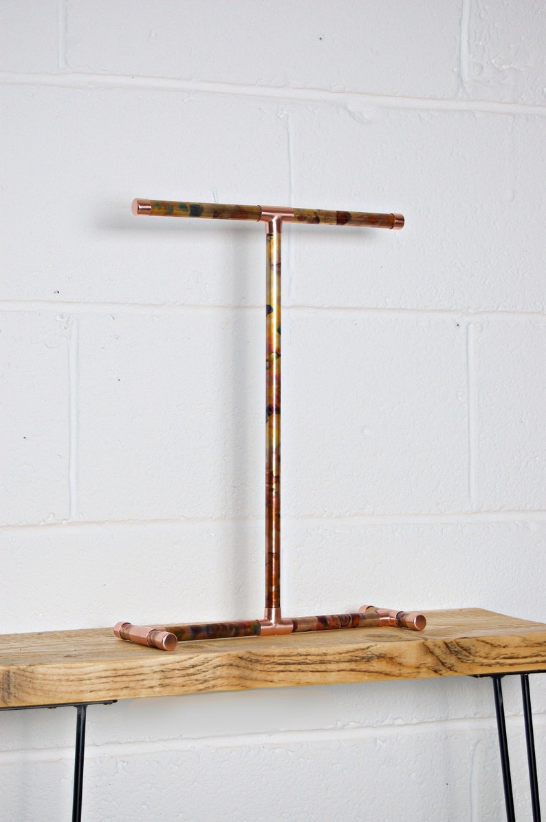 May include: A copper jewelry stand with a T-shaped top bar and a base with three legs. The stand is made of copper pipe and has a rustic, industrial look.