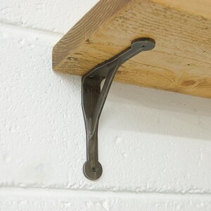 A solid cast iron shelf bracket on a white painted brick wall, with a rustic wooden shelf.
