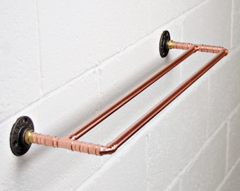 May include: Copper towel rack with two bars mounted on a white brick wall. The rack has black metal flanges on each end.