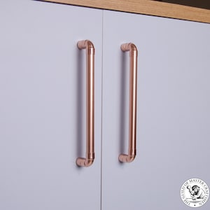 Könnte beinhalten: Zwei Schranktürgriffe aus Kupferrohr, montiert an einer hellgrauen Schranktür. Die Griffe sind warm, rötlich-braun und haben ein gebogenes Design. Die Schranktür ist Teil eines größeren Schranks mit einer Holzmaserungsoberseite.
