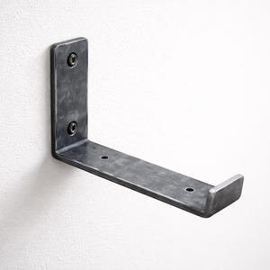 Peut inclure: Un support d'étagère en métal gris foncé en forme de L, monté sur un mur blanc. Le support est en métal épais et comporte deux vis sur le côté vertical pour le montage. Le côté horizontal a deux trous et un petit rebord.
