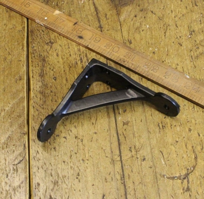 A small 4" solid cast iron shelf bracket finished in black on a wooden floorboard surface with a ruler at the top for a guide on size.