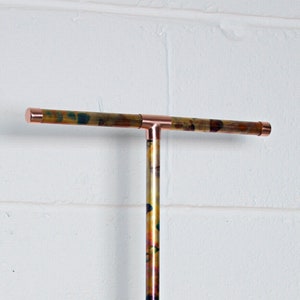 May include: A copper T-shaped pipe with a mottled, iridescent finish. The pipe is mounted to a white wall.