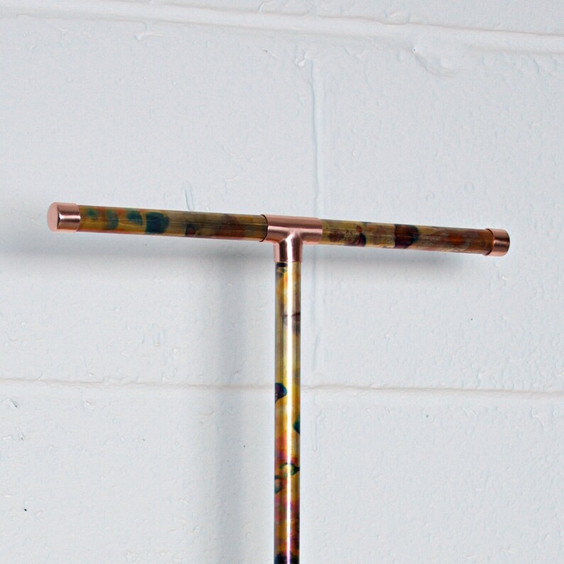 May include: A copper T-shaped pipe with a mottled, iridescent finish. The pipe is mounted to a white wall.