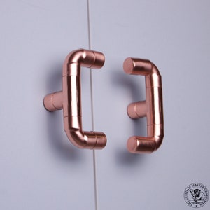 Puede incluir: Dos tiradores de puerta de armario de estilo tubería de cobre. Los tiradores tienen forma de letra 'E' y están montados en una superficie blanca.