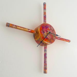 May include: A copper clock with a circular face and four arms extending out from the centre. The clock face has a marbled pattern of red, orange, and yellow colours. The arms are also copper and have a marbled pattern.