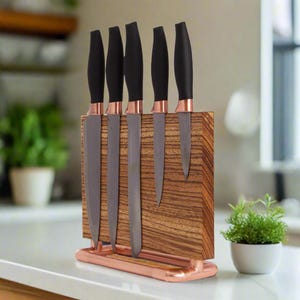 Op de afbeelding: Een set van zes keukenmessen met zwarte handvatten en koperkleurige bolster, gepresenteerd in een houten blok met een koperkleurige basis. De messen hebben zilveren bladen en zijn in een rij gerangschikt. Het houten blok heeft een streepjespatroon.