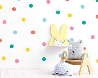 May include: A white wall with colorful polka dot stickers. A yellow bunny rabbit shelf with a clock and a gray and white stuffed animal sitting on a wooden chair with a blue whale stuffed animal on the floor.