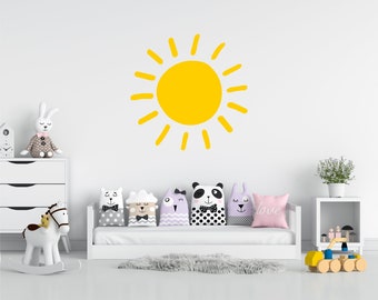 May include: A white rocking horse with a yellow sun decal on the wall behind it. The horse is in a nursery with a white couch and pillows with animal faces on them.