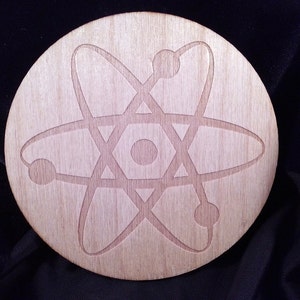 May include: A round wooden coaster with a laser-etched design of an atom. The atom is depicted with a central nucleus and orbiting electrons.