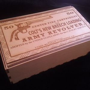 May include: A wooden box with an engraved design. The box is labeled "50 Center Fire Cartridges for Colt's New Breech Loading Army Revolver." The box is made by the Union Metallic Cartridge Co.
