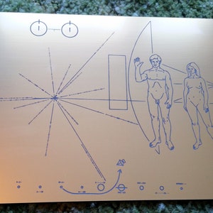 May include: A gold-colored metal plaque with a blue line drawing of a stylized human figure, a circle with a line through it, and a series of lines radiating from a central point. The plaque also includes a diagram of a solar system with a circle representing the sun and smaller circles representing planets.