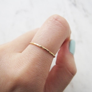 May include: A delicate, hammered gold ring. The thin band has a textured surface and is worn on a finger. The ring's warm gold colour contrasts with the skin tone and the light background. A simple, elegant piece of jewellery.
