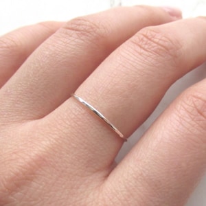 May include: A slender silver ring is worn on a finger. The ring has a minimalist design, appearing to be a single, thin band. The ring's polished surface reflects light, creating a subtle shine. The ring is a piece of jewellery. The ring's band is approximately 1 mm thick.