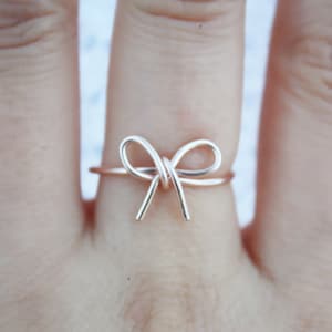 May include: A delicate gold-toned ring featuring a simple wire bow design. The ring is worn on a finger.
