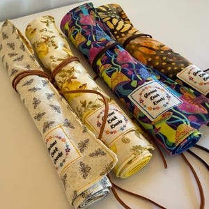 May include: Rolled fabric cases in various patterns, including bees, floral, and mushroom designs. Each case is secured with a brown leather cord. The cases have a label that reads "Glorious Knick Knacks".