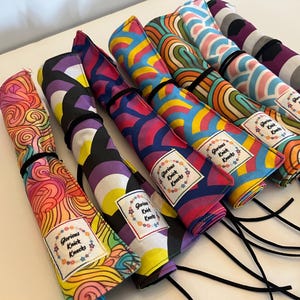 May include: A collection of colorful fabric rolls with various patterns, including swirls, rainbows, and geometric designs. Each roll is secured with a black elastic band and features a label that reads "Glorious Knick Knacks."