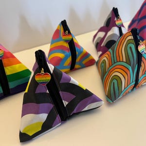 May include: A collection of triangular pouches with various colorful patterns, including rainbow stripes, geometric designs, and abstract art. Each pouch has a black zipper and a heart-shaped charm with a rainbow design.
