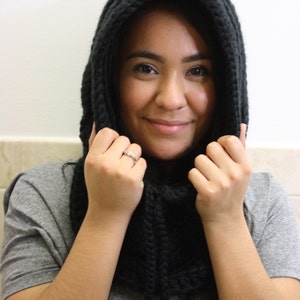 May include: A woman wearing a black knitted hooded scarf. The scarf is pulled up over her head and she is holding the edges of the scarf with both hands.