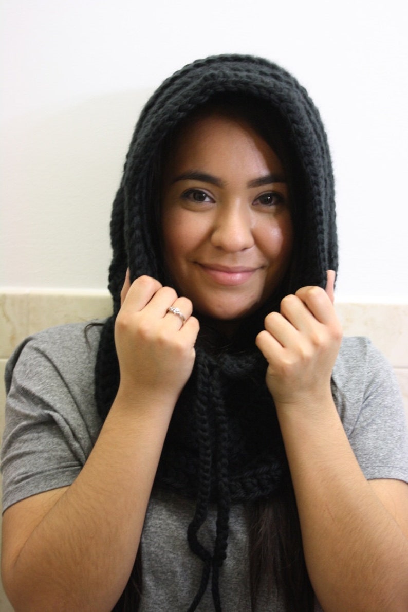 May include: A woman wearing a black knitted hooded scarf. The scarf is pulled up over her head and she is holding the edges of the scarf with both hands.