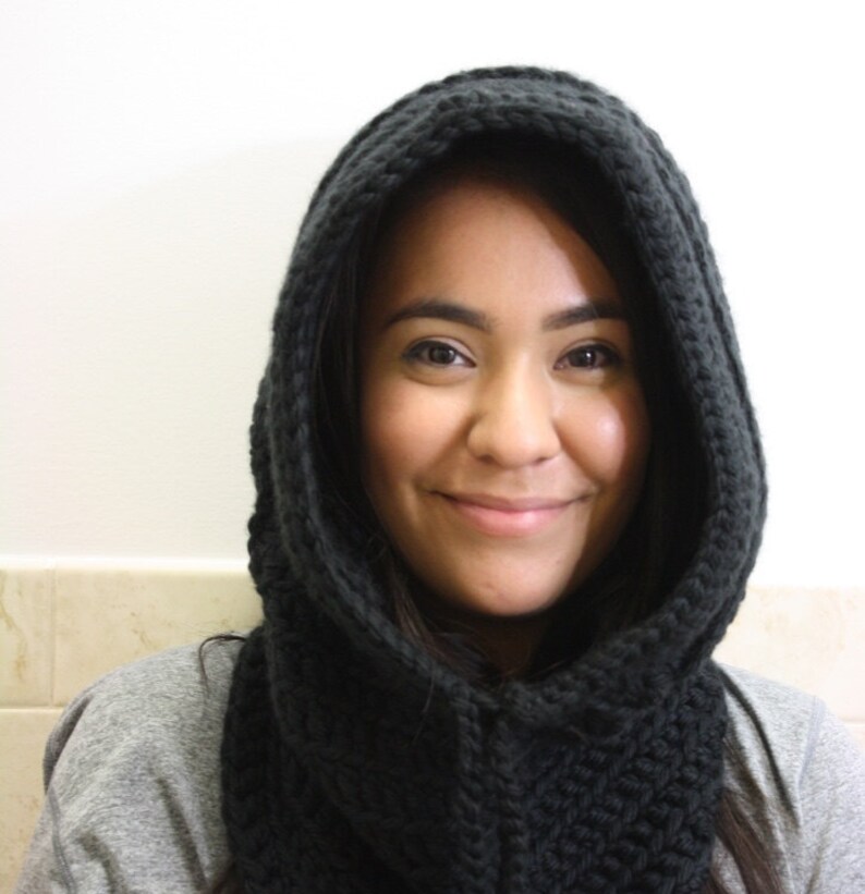 May include: A woman wearing a black knitted hooded scarf. The scarf is thick and has a cable knit pattern.