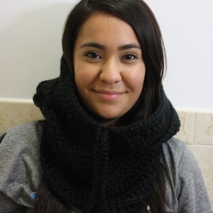 May include: A woman with long dark hair is wearing a black crocheted cowl neck scarf. The scarf is thick and has a textured pattern. She is wearing a gray shirt.