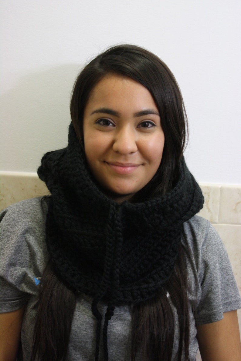 May include: A woman with long dark hair is wearing a black crocheted cowl neck scarf. The scarf is thick and has a textured pattern. She is wearing a gray shirt.