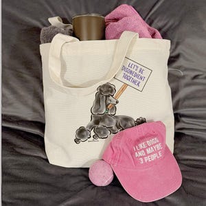 May include: A cream-colored tote bag with a poodle graphic holding a sign that reads "LET'S BE DISOBEDIENT TOGETHER." A pink baseball cap with the text "I LIKE DOGS AND MAYBE 3 PEOPLE" and a pink tennis ball are next to the bag.
