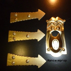 May include: Three gold-colored metal arrow-shaped door hinges and a gold-colored metal door handle with a face and keyhole. The door handle has the text "Mancaveprop" on the bottom.