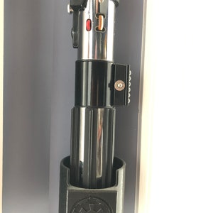May include: A black lightsaber holder with a silver lightsaber inside. The holder has a black and white Star Wars logo on the front.