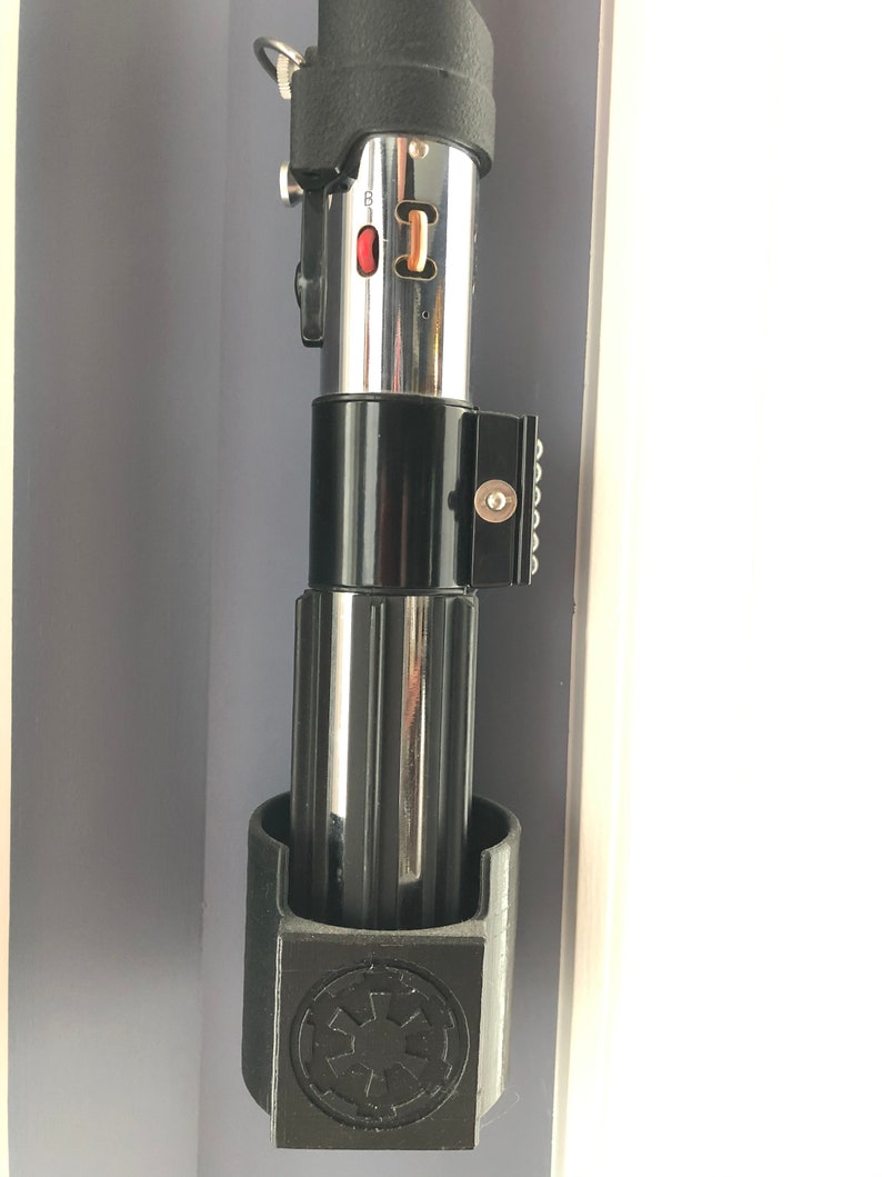 May include: A black lightsaber holder with a silver lightsaber inside. The holder has a black and white Star Wars logo on the front.