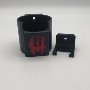May include: A black 3D printed wall mount with a red logo. The mount has a cylindrical cup for holding an object and a separate bracket for attaching to a wall.