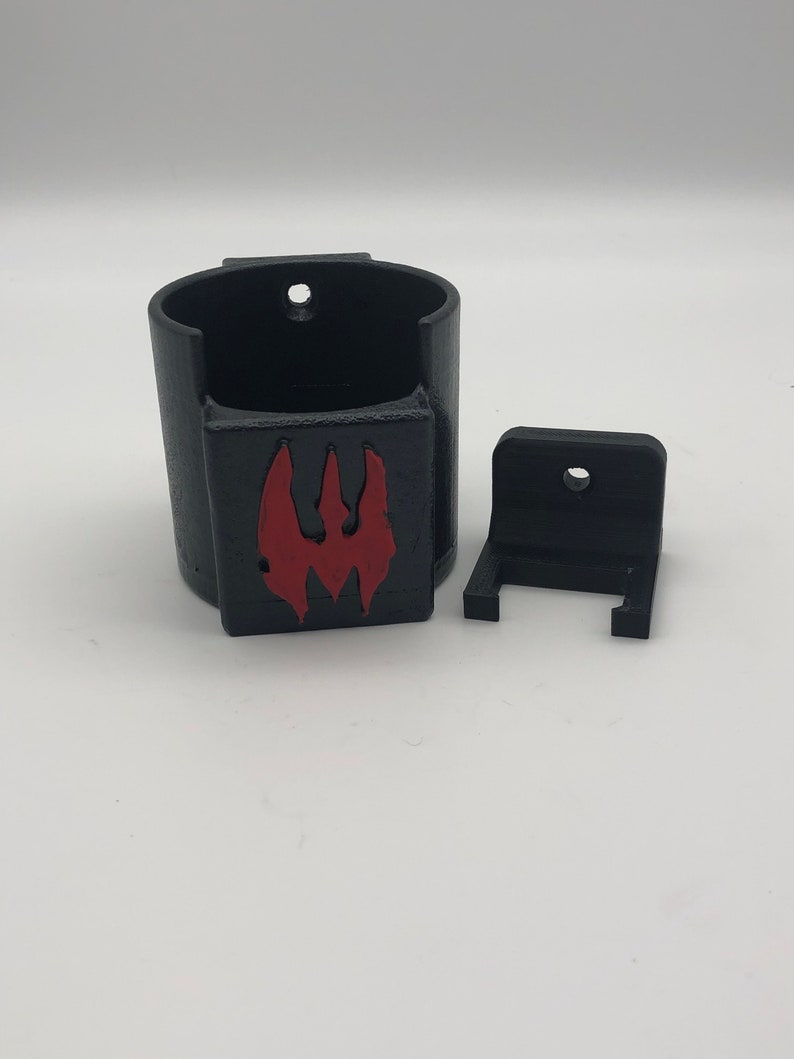 May include: A black 3D printed wall mount with a red logo. The mount has a cylindrical cup for holding an object and a separate bracket for attaching to a wall.