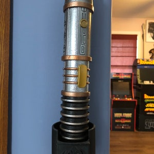 May include: A silver and gold lightsaber with a black lightsaber holder featuring the Rebel Alliance symbol. The lightsaber has a textured surface and a gold band around the middle.