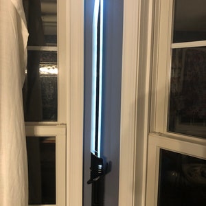 May include: A black lightsaber with a white blade is mounted on a wall. The lightsaber has a black handle with a red symbol on the bottom.