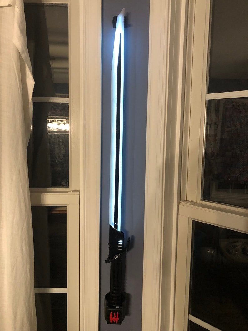 May include: A black lightsaber with a white blade is mounted on a wall. The lightsaber has a black handle with a red symbol on the bottom.