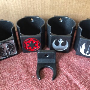May include: Four black wall-mounted cup holders with different Star Wars logos. The logos are the Rebel Alliance symbol, the Imperial symbol, the Death Star symbol, and the X-Wing symbol. The holders are made of black plastic and have a textured finish.