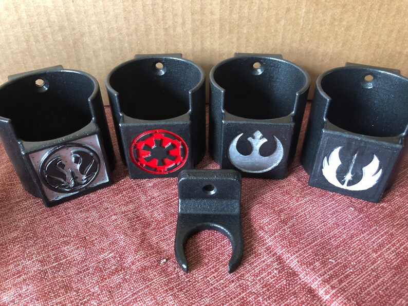 May include: Four black wall-mounted cup holders with different Star Wars logos. The logos are the Rebel Alliance symbol, the Imperial symbol, the Death Star symbol, and the X-Wing symbol. The holders are made of black plastic and have a textured finish.