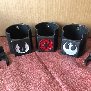 May include: Three black wall-mounted cup holders with different Star Wars logos. The logos are the Rebel Alliance symbol, the Imperial symbol, and the symbol of the Mandalorian faction. Each holder has a small hook on the side.